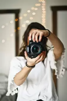A woman holds a camera to the viewer against a blurred background