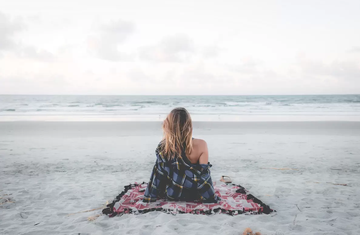 Picture of a person sitting alone on a towel at an empty beach. She is facing the water.